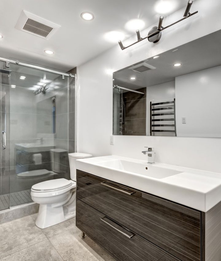 Stylish bathroom featuring a large, frameless glass-enclosed shower with dark grey tiles. The space includes a modern vanity with a long white sink and dark wood cabinets, complemented by a large mirror and minimalist lighting. The overall design is clean and contemporary, emphasizing functionality and modern aesthetics.
