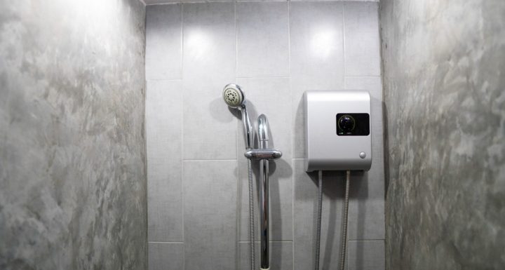 Modern compact shower cubicle featuring grey textured tile walls with a streamlined design. Equipped with a handheld shower head and an innovative wall-mounted digital water heater unit for instant temperature control, this setup optimizes space and functionality for a practical bathroom solution.