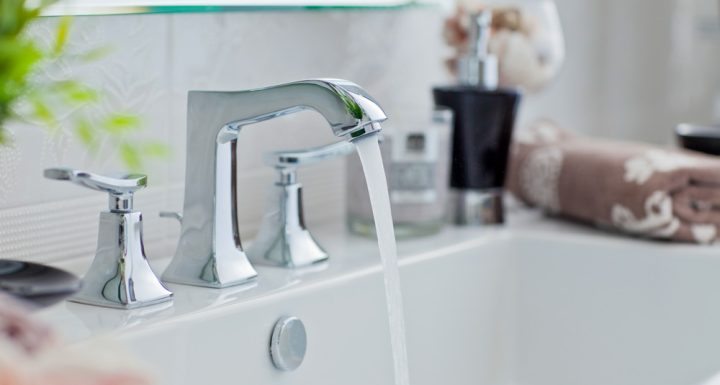 Close-up of a bathroom sink featuring sleek chrome faucets with a modern design. Water is flowing from the tap, adding a dynamic element to the clean and sophisticated setup. The background includes a blurred view of bathroom accessories and a neatly folded towel, enhancing the elegant and functional ambiance.